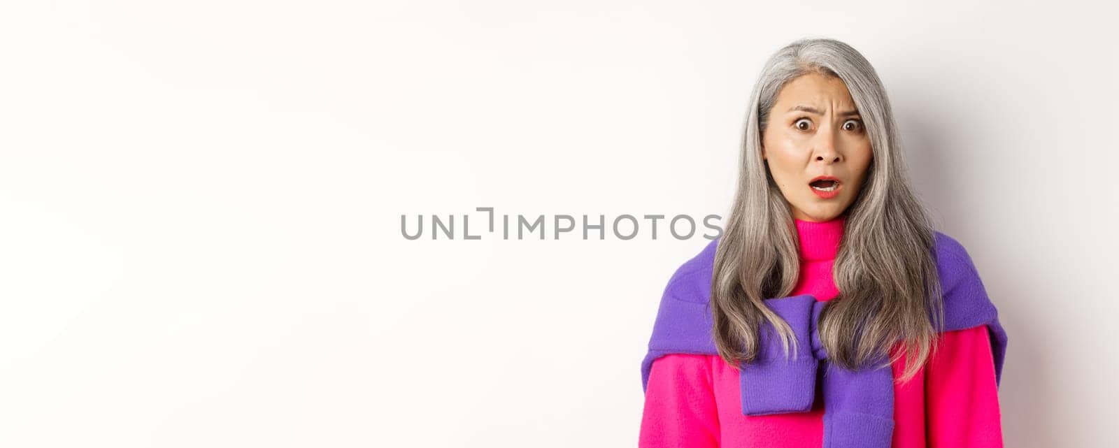 Close-up of shocked asian senior woman gasping, staring with confused face, looking with cringe, standing against white background.