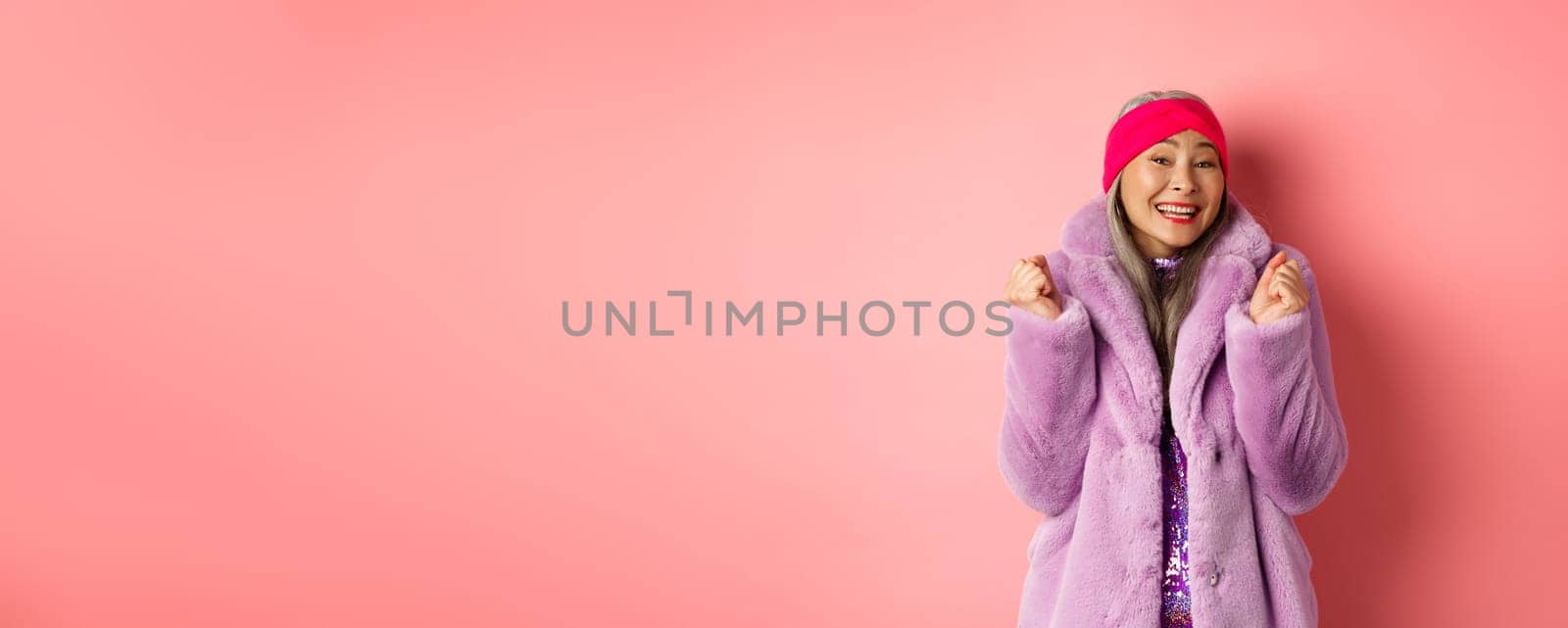 Cheerful asian lady in stylish funky coat, celebrating victory or success, saying yes and smiling happy, standing over pink background by Benzoix