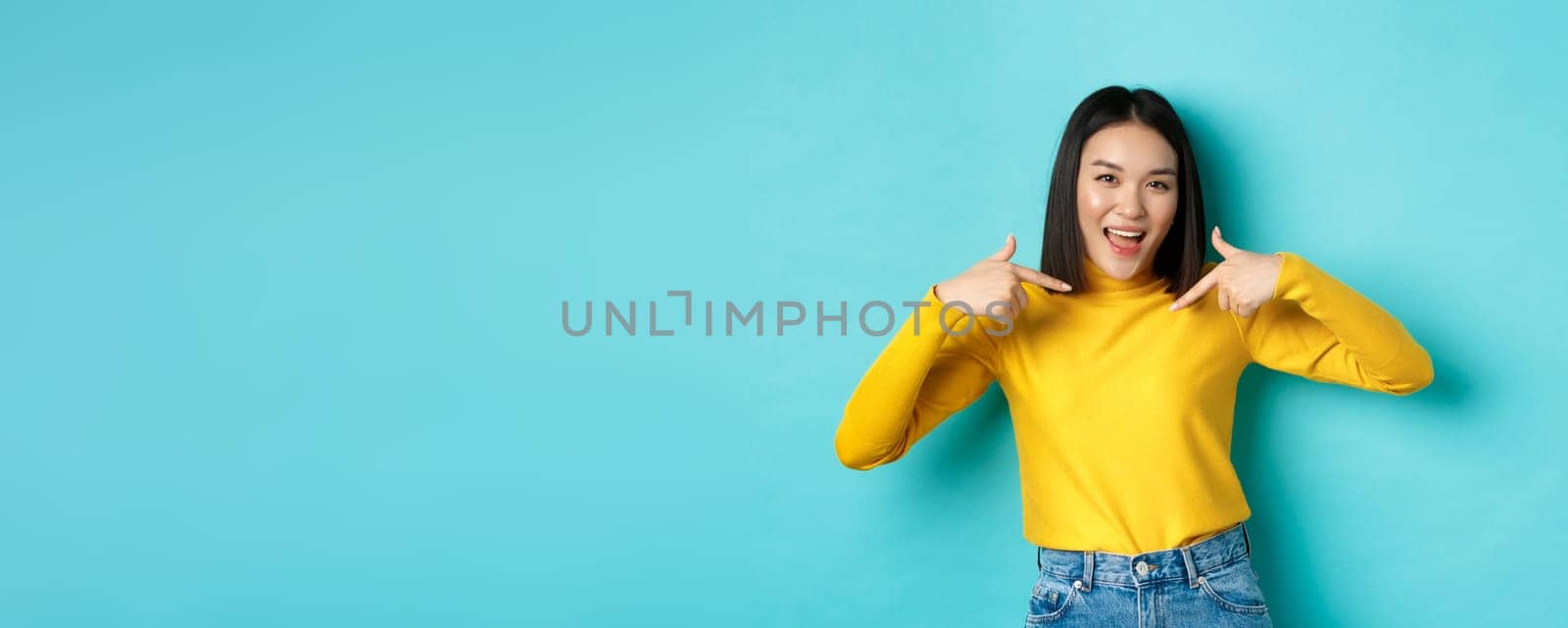 Beauty and fashion concept. Confident and sassy asian woman pointing at herself, smiling cheeky at camera, being professional, standing over blue background.