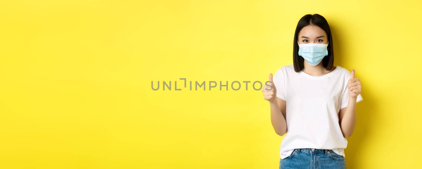 Covid-19, quarantine and social distancing concept. Cheerful asian woman in medical mask and white t-shirt showing thumbs up in approval, praise good deal, yellow background.