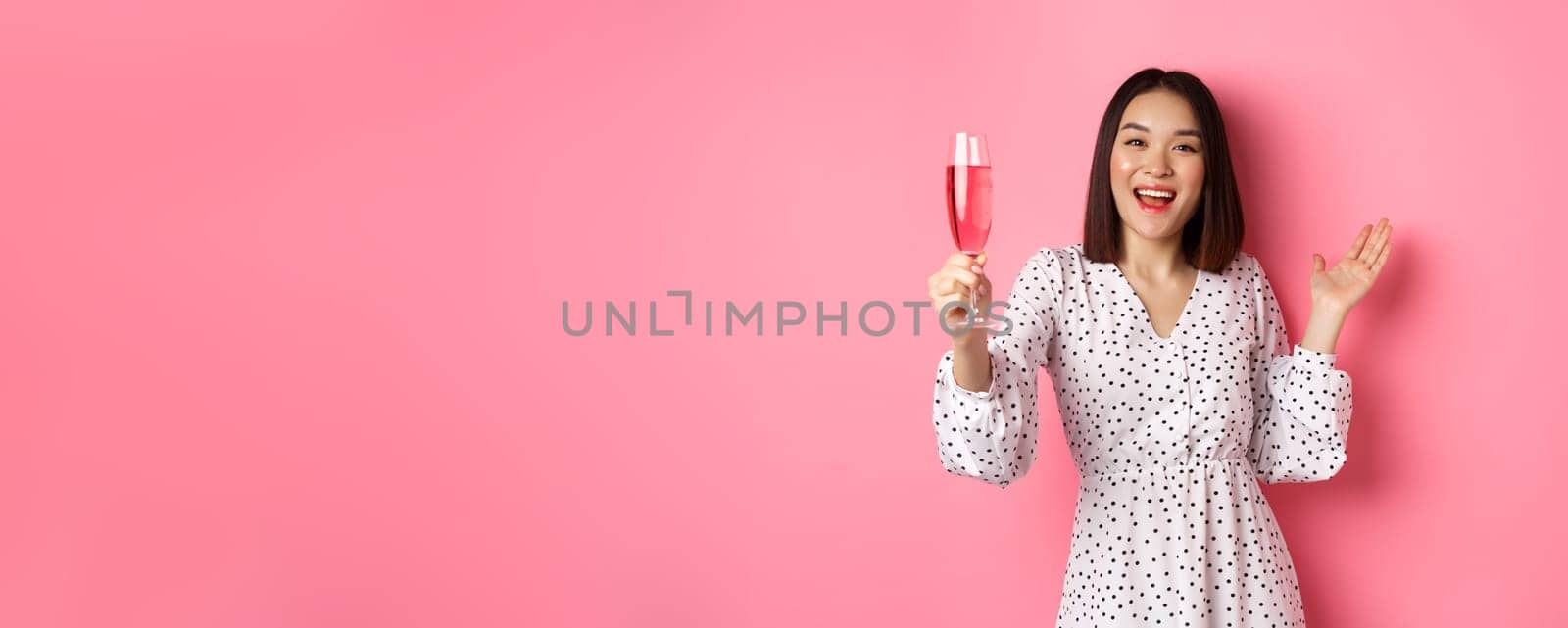 Happy asian woman celebrating, saying toast on party, raising glass of champagne and smiling, standing in dress over pink background.