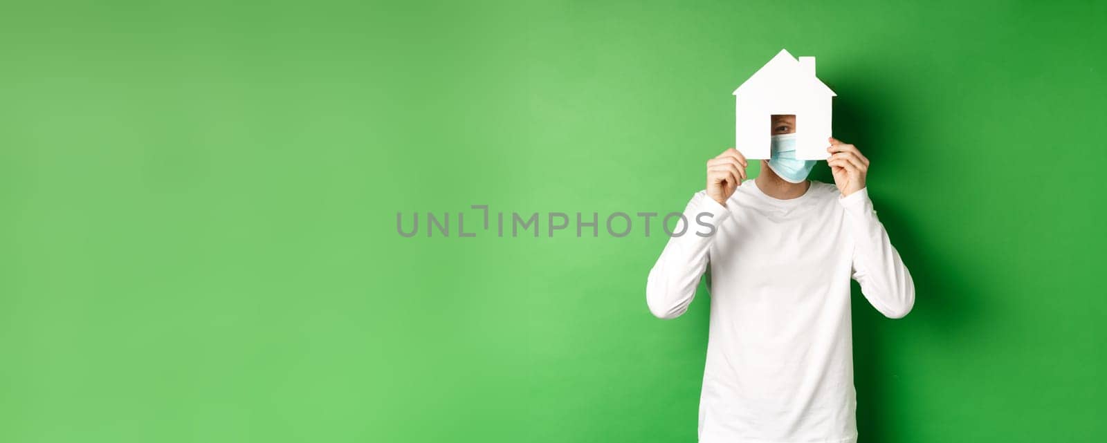 Real estate and covid-19 pandemic concept. Funny young man in face mask and white long-sleeve hiding face behind paper house cutout, peeking at camera, green background.