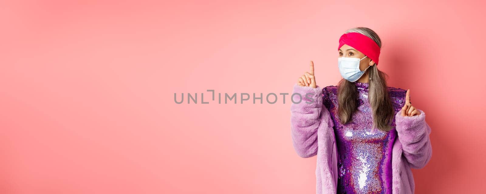 Covid-19, social distancing and fashion concept. Fancy asian senior woman dancing and having fun, wearing medical respirator and purple shiny dress, pink background by Benzoix