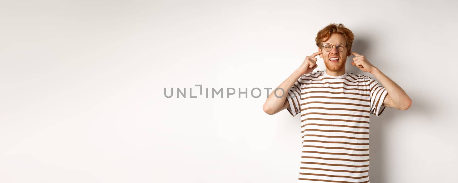 Annoyed redhead guy in glasses shutting ears and complaining loud noise, angry at noisy neighbours, standing over white background by Benzoix