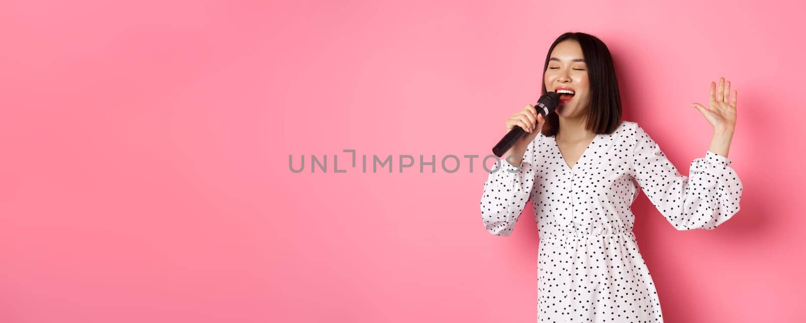 Happy asian woman dancing and singing in microphone, performing at karaoke, standing over pink background by Benzoix