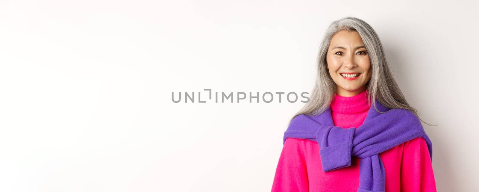 Close-up of beautiful asian senior woman in trendy pink sweater, smiling cheerful at camera, standing over white background.
