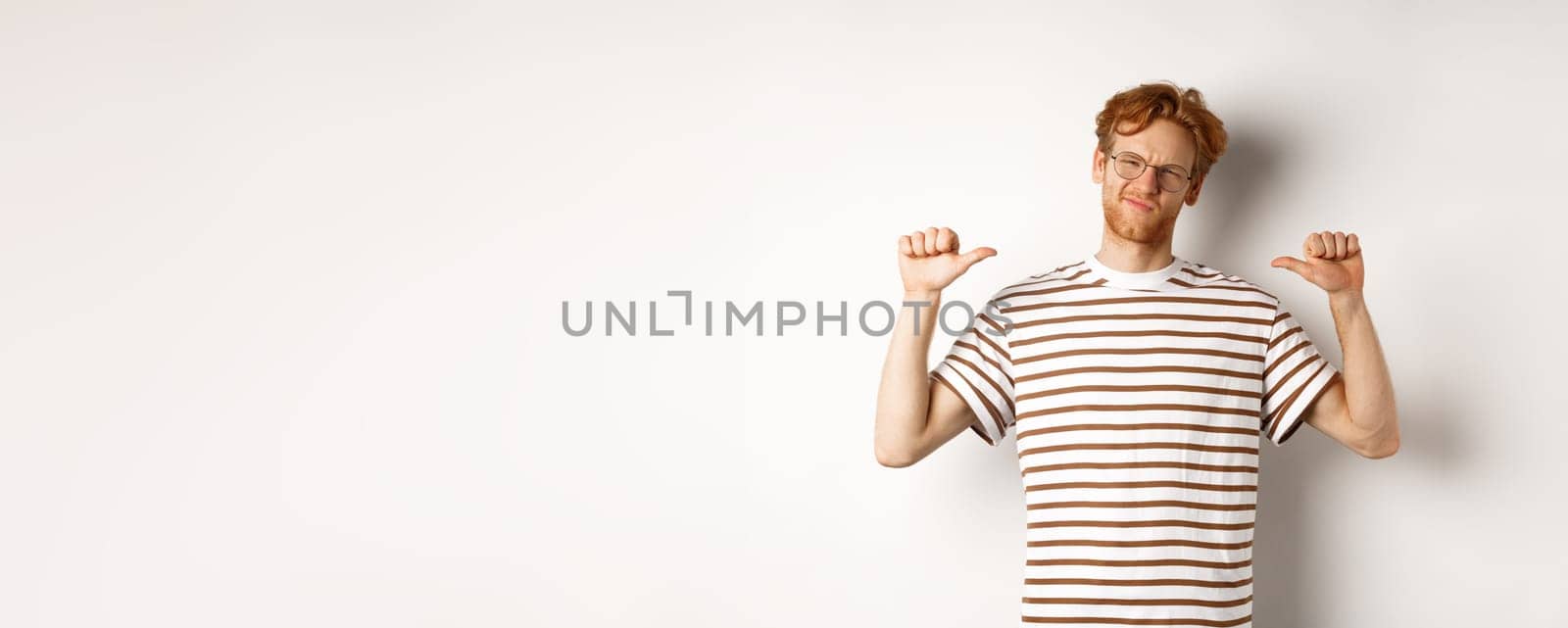 Confident young nerdy guy in glasses with red hair, pointing at himself and looking like professional, white background.