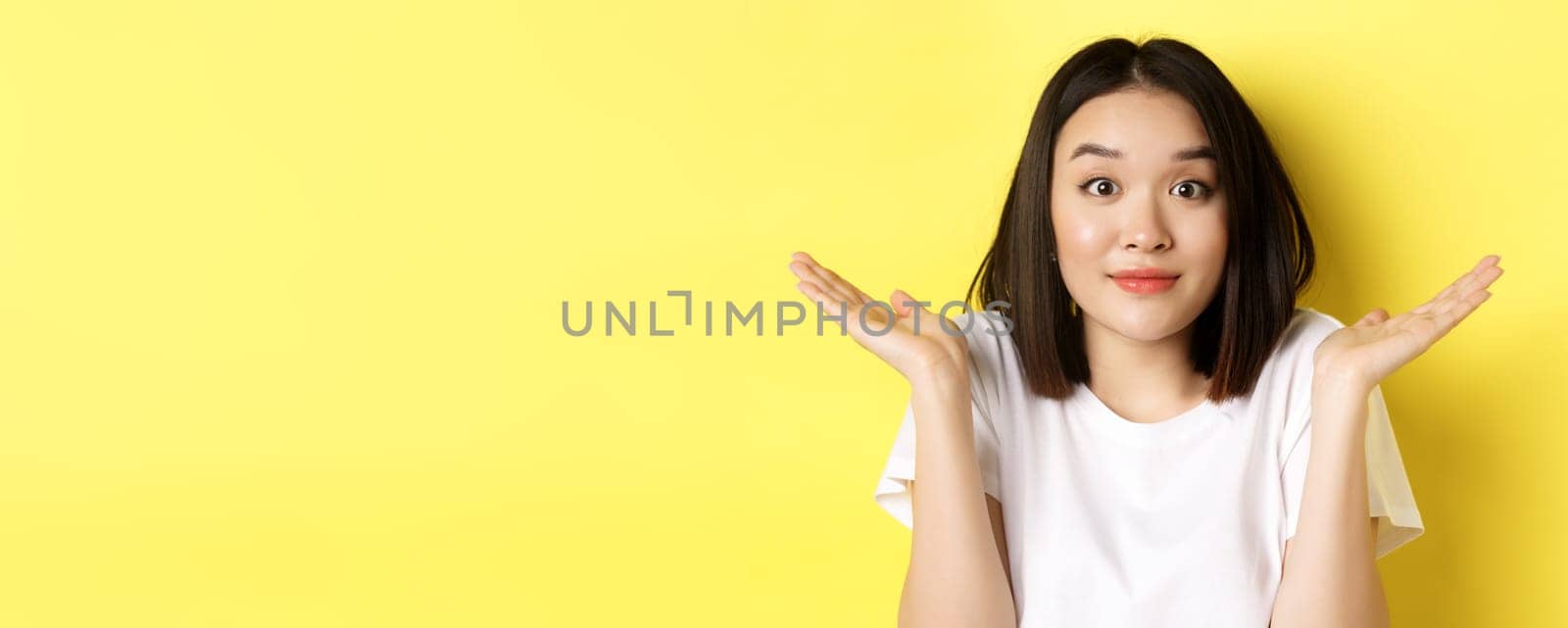 Silly mistake. Close up of cute asian girl saying sorry, shrugging shoulders and smiling with oops face expression, standing over yellow background by Benzoix