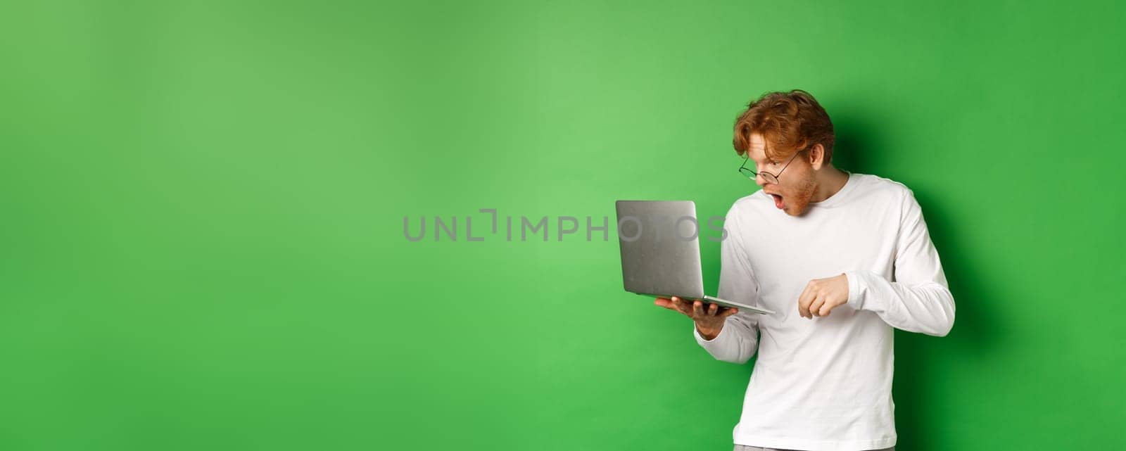 Image of young redhead man looking surprised at laptop screen, drop jaw and staring at display amazed, green background by Benzoix