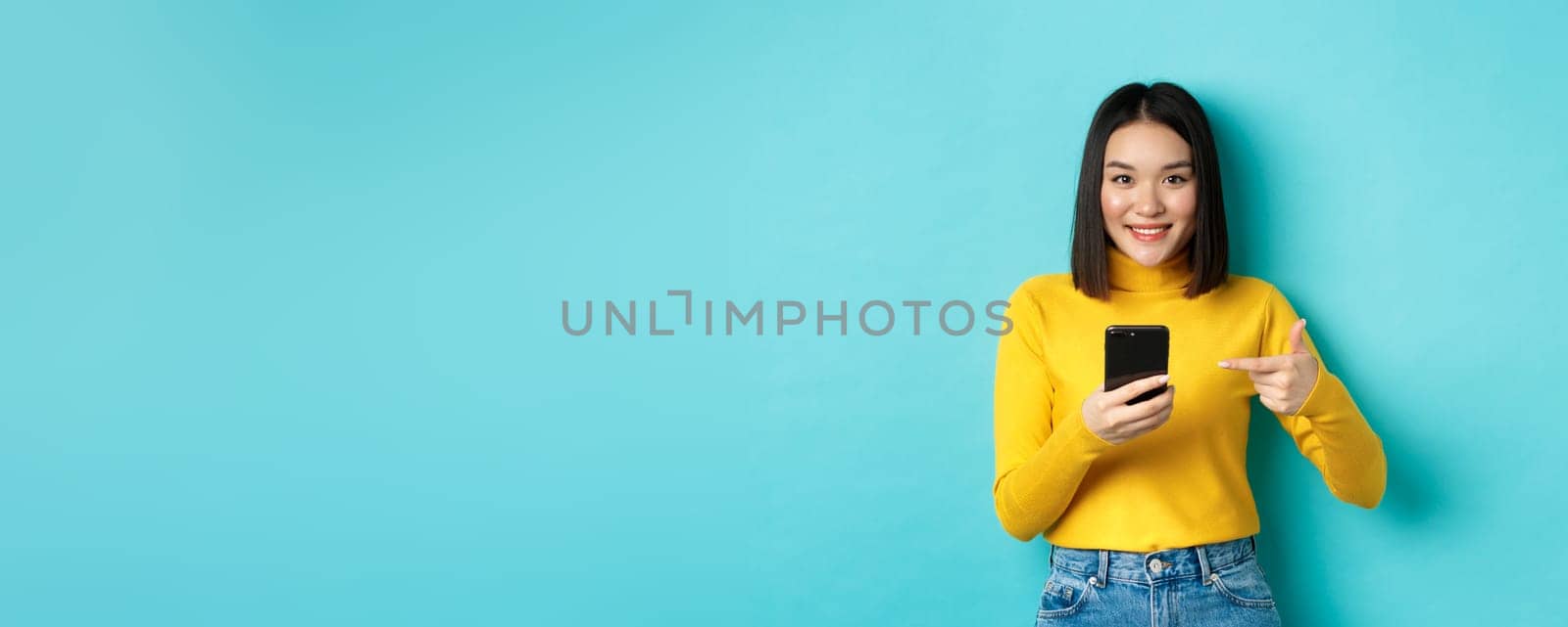 E-commerce and online shopping concept. Cute asian woman in yellow sweater pointing at smartphone, smiling at camera, standing over blue background by Benzoix