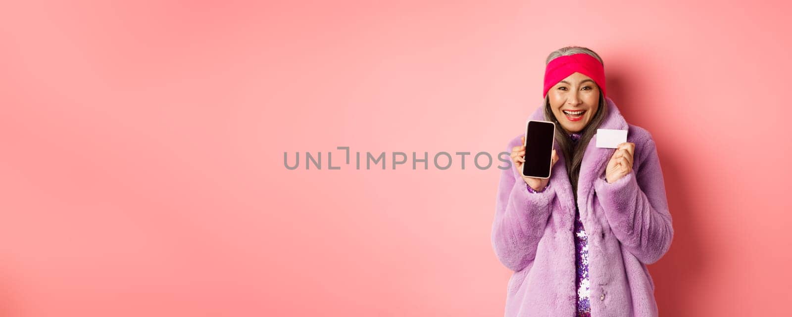 Online shopping and fashion concept. Beautiful asian mature woman showing plastic credit card and blank phone screen, smiling happy, pink background.