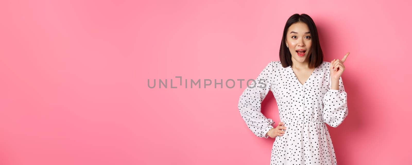 Excited pretty asian girl pointing at upper right corner, gasping amazed and looking at camera, discuss store promo offer, standing over pink background.