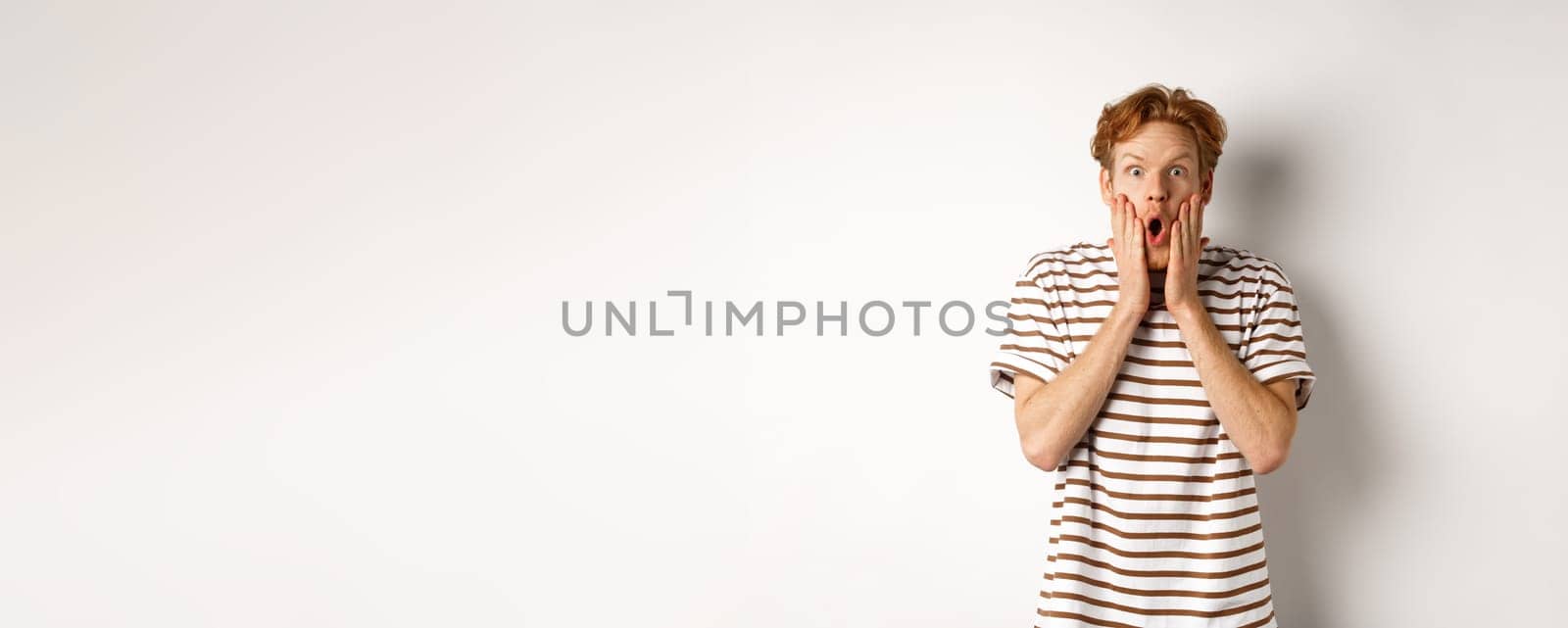 Impressed young man with ginger hair, gasping and staring shocked at camera, express complete disbelief and amazement, white background.