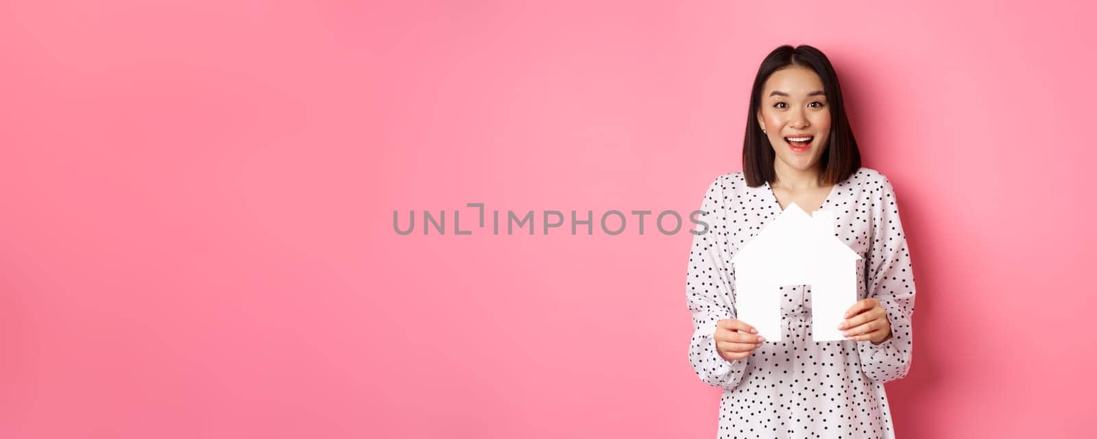 Real estate. Amazed asian woman searching for flat, holding house model and looking at camera happy, standing over pink background by Benzoix