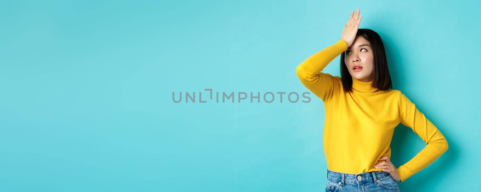Emotions and lifestyle concept. Annoyed asian girl roll eyes and face palm, standing bothered in yellow pullover against blue background.