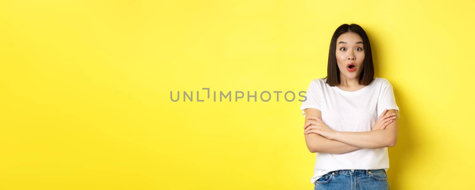 Portrait of surprised asian girl checking out promo, gasping amazed, staring at camera amazed, yellow background.