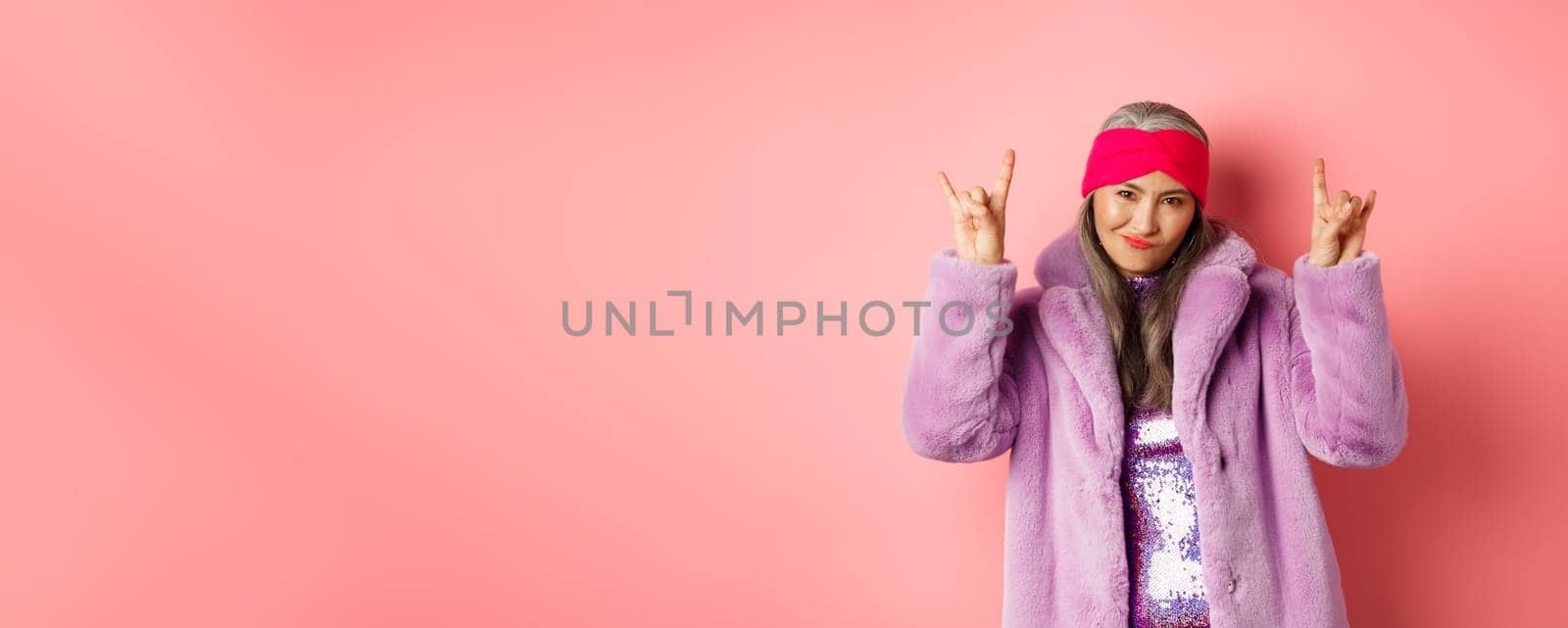 Cool asian mature woman in fashionable faux fur coat, showing rock-n-roll horns signs and smiling sassy at camera, having fun, standing over pink background.