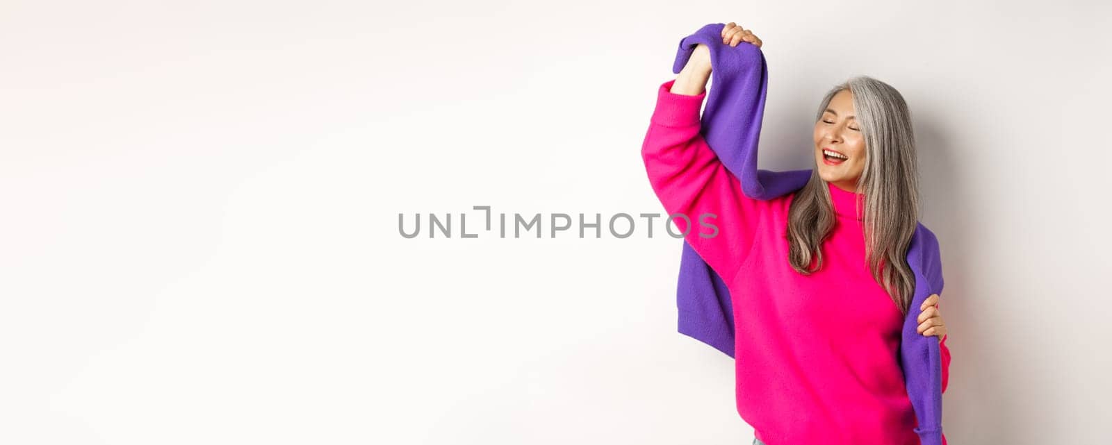 Carefree korean elderly woman in pink sweater, dancing with sweatshirt on shoulders and smiling, posing happy over white background.