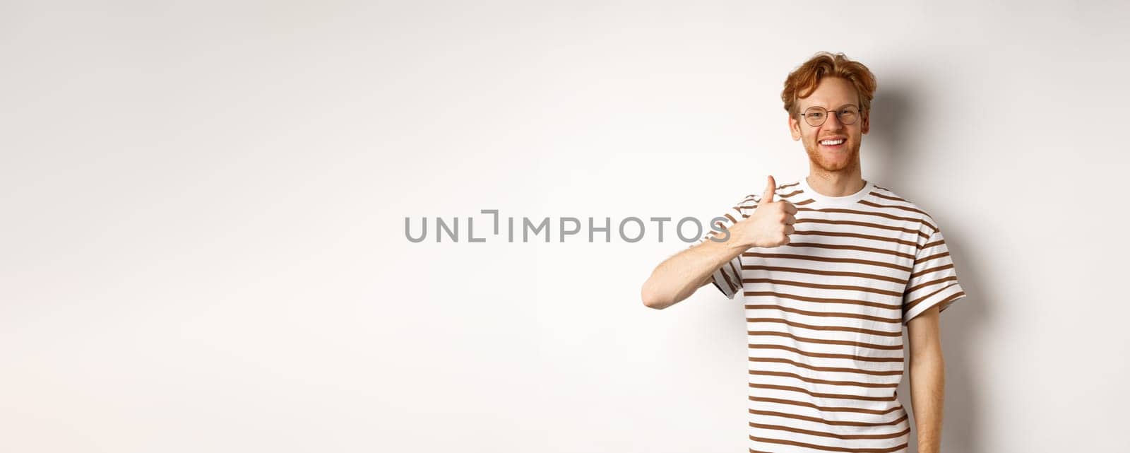 Smiling young male student with red hair and bristle, showing thumb-up and looking satisfied, leave positive feedback, recommending promo, white background.