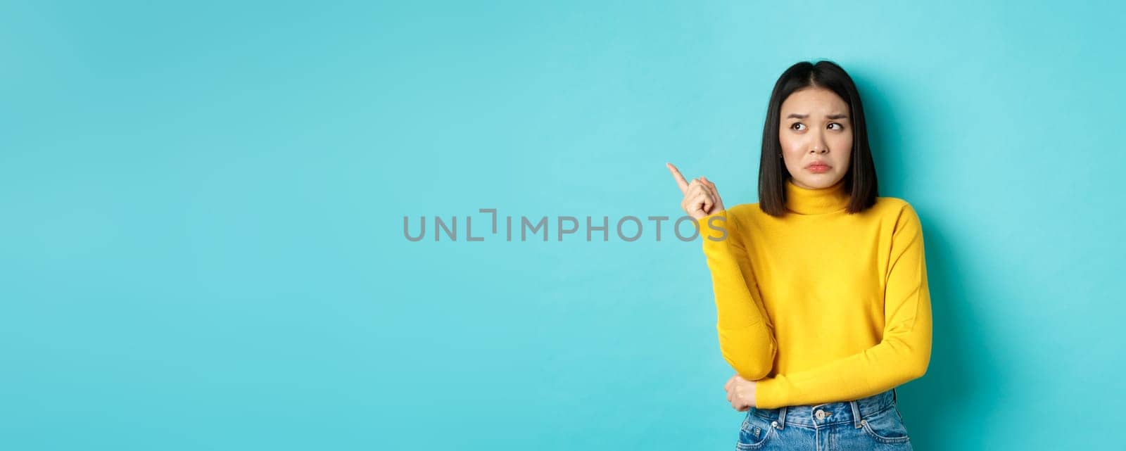 Shopping concept. Disappointed and gloomy asian woman sulking upset, pointing finger left at bad news banner, standing over blue background by Benzoix