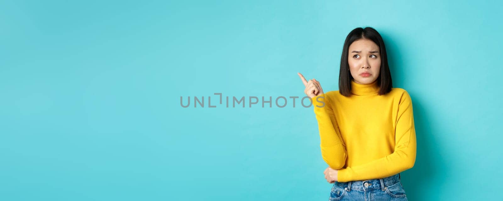 Shopping concept. Disappointed and gloomy asian woman sulking upset, pointing finger left at bad news banner, standing over blue background.