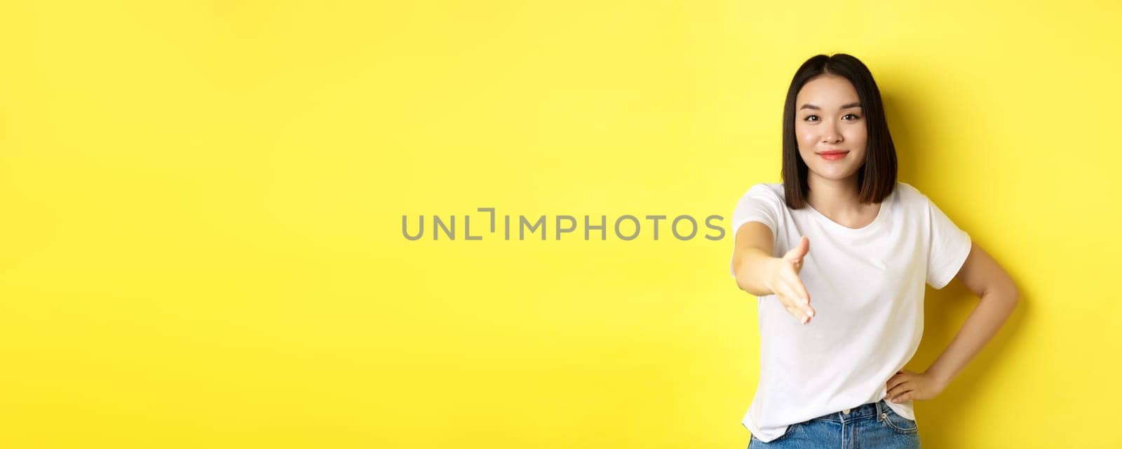 Confident asian woman in white t-shirt, stretch out hand for handshake and greeting gesture, saying hello, introduce herself, standing over yellow background.