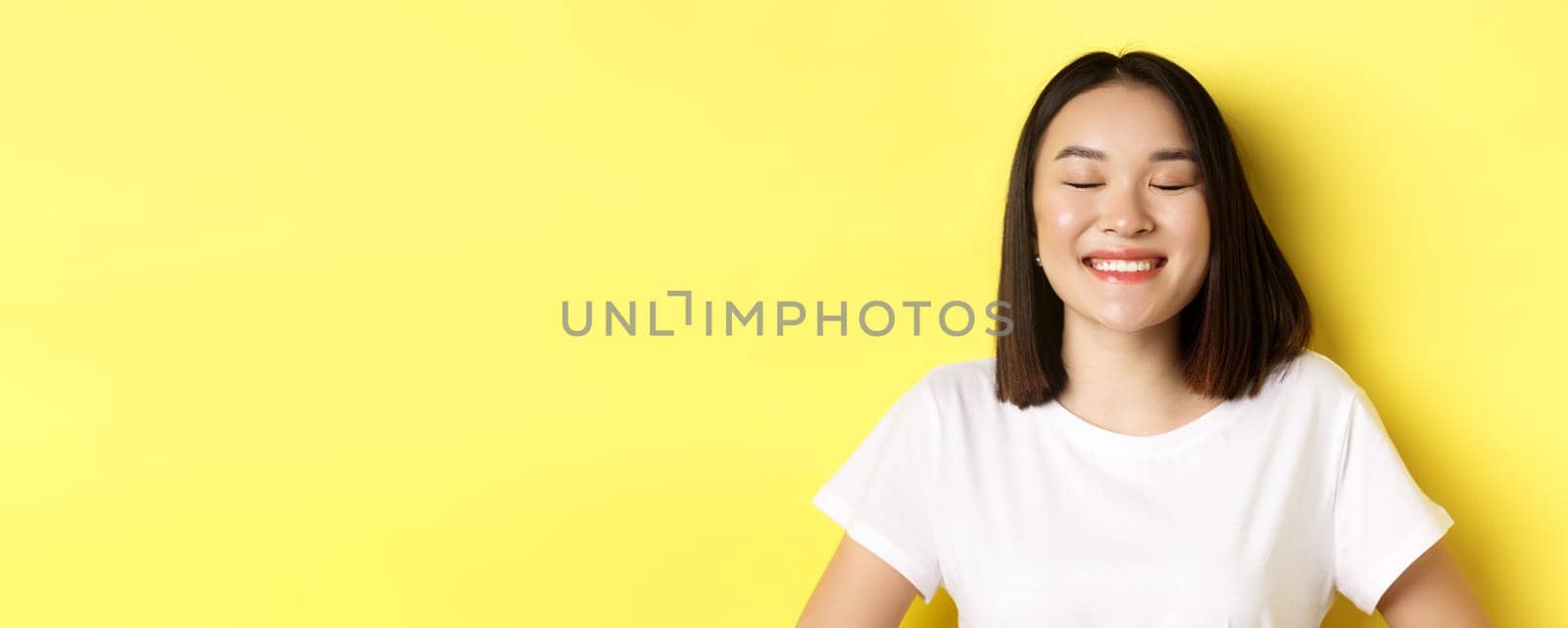 Close up of happy, romantic asian girl dreaming of something, close eyes and smiling delighted, standing over yellow background by Benzoix