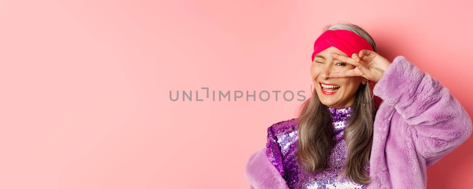 Fashion. Close-up of fashionable asian senior woman smiling, showing victory sign over eye and looking happy at camera, standing over pink background.