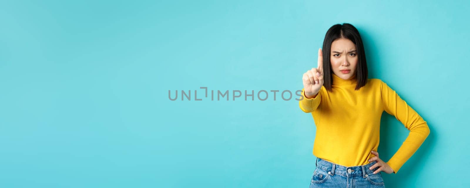Confident and serious woman tell no, showing extended finger to stop and prohibit something bad, frowning and looking at camera self-assured, standing over blue background.