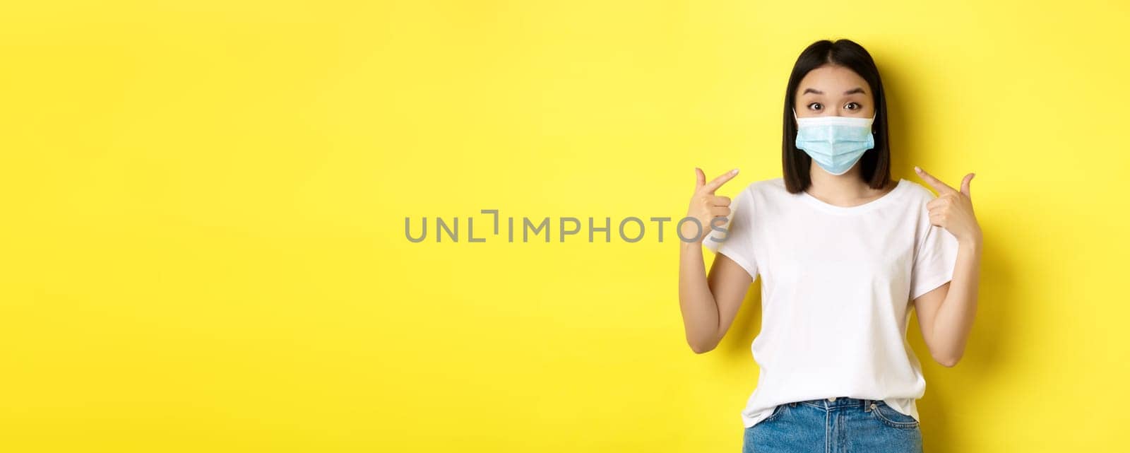 Covid-19, quarantine and social distancing concept. Young asian woman in white t-shirt, pointing at her medical mask from coronavirus, yellow background by Benzoix