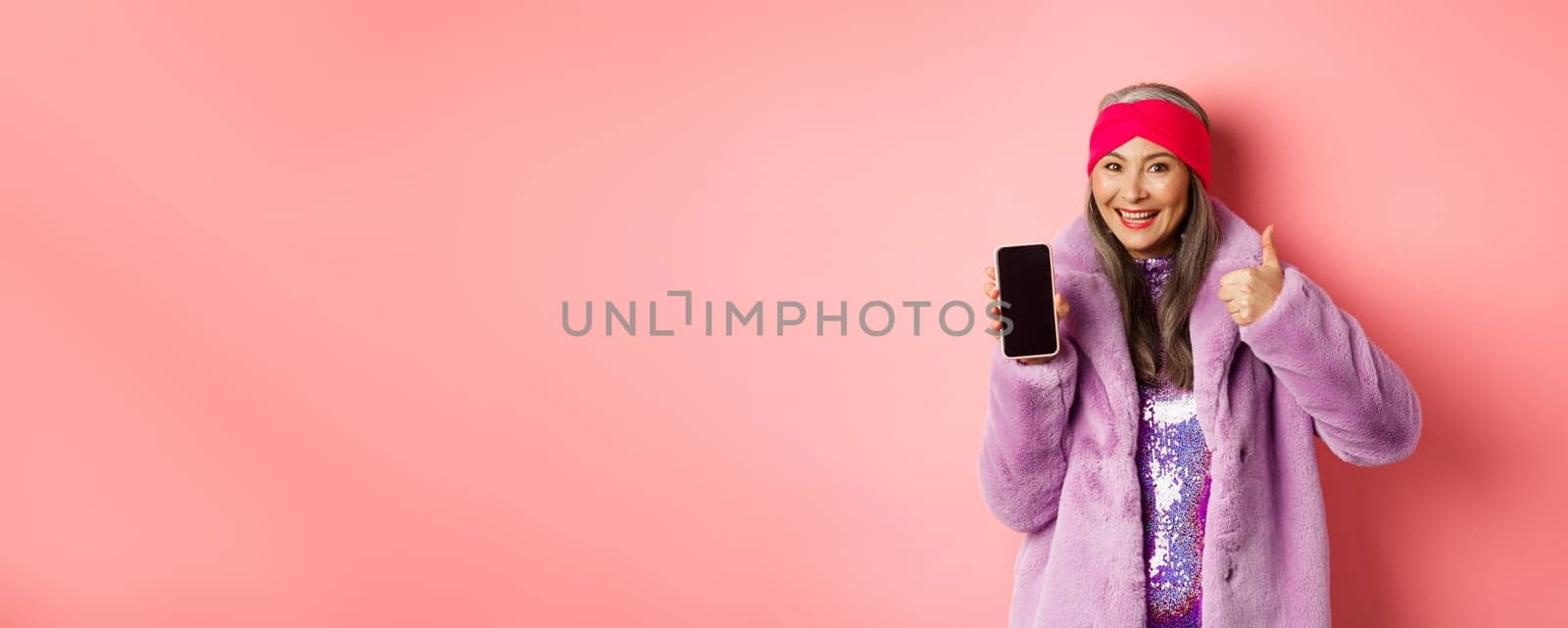 Online shopping and fashion concept. Funky asian senior woman showing blank mobile screen and thumbs-up, like and recommend internet promo, pink background.