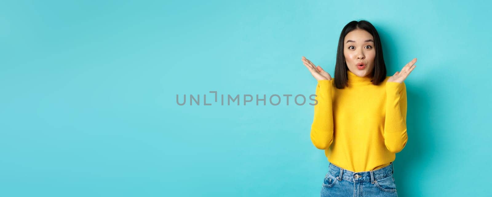Beauty and fashion concept. Image of excited and surprised japanese girl saying wow with amazement, raising hands up near face, standing against blue background.