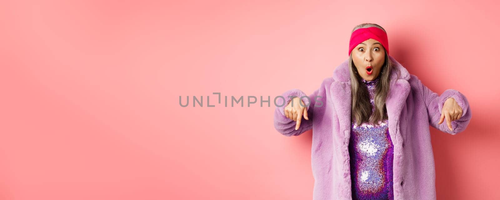 Fashion and shopping concept. Impressed asian female pointing fingers down and looking excited at camera, showing awesome promo offer, standing over pink background.