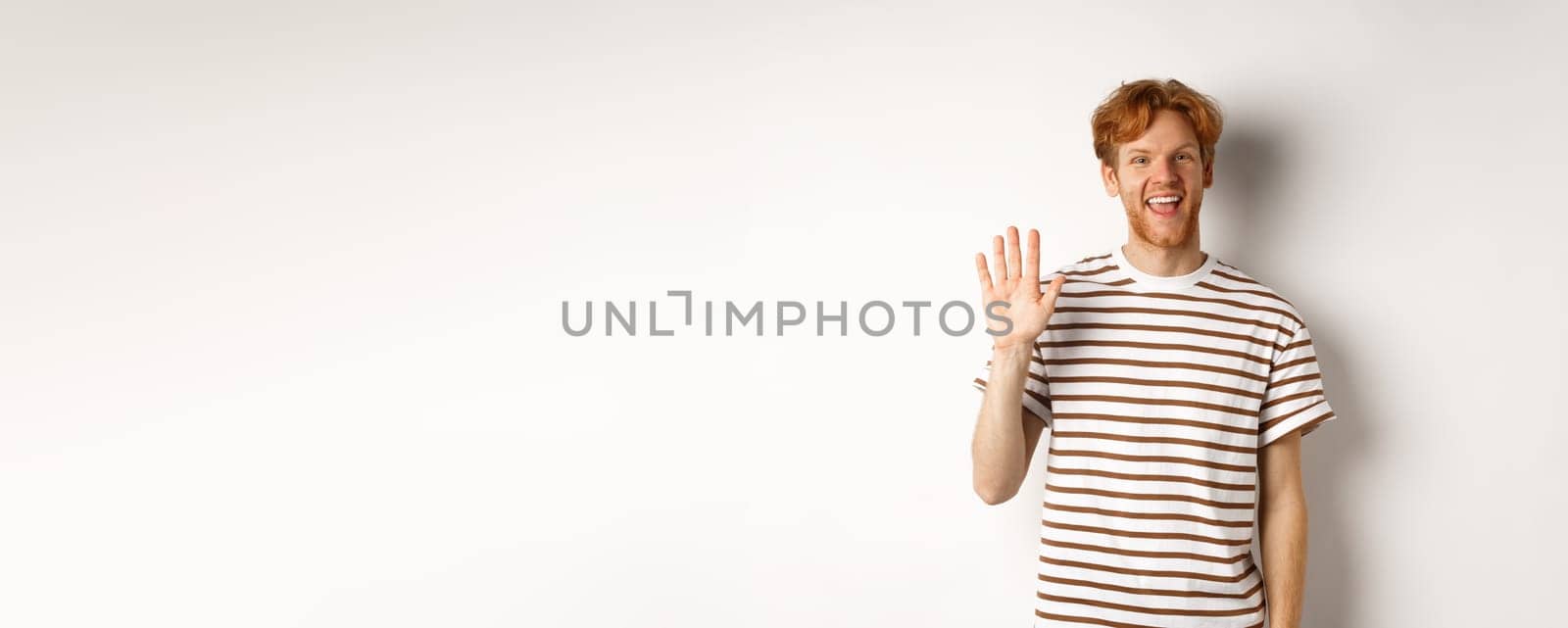 Friendly man with red hair and beard saying hi, waving hand and smiling, standing over white background.