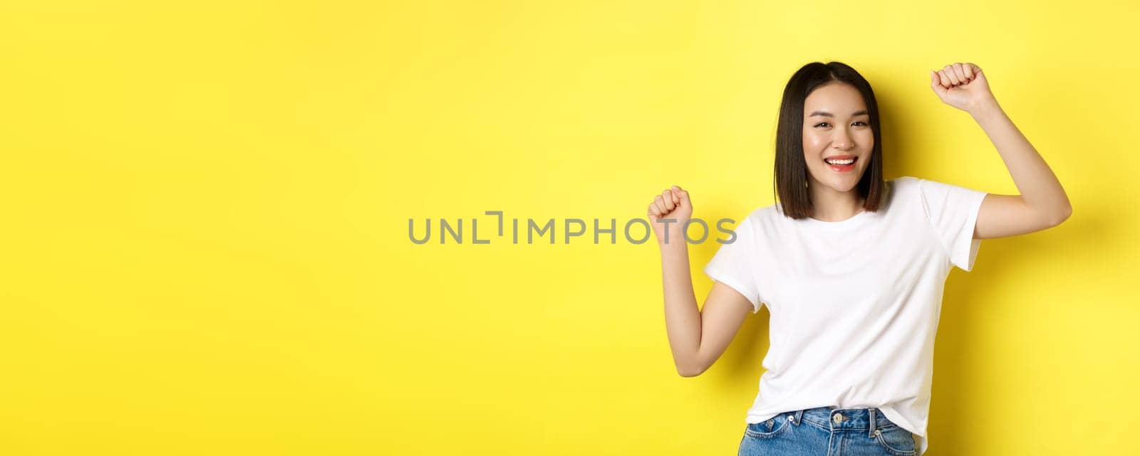 Carefree asian girl dancing and having fun, posing in white t-shirt against yellow background by Benzoix