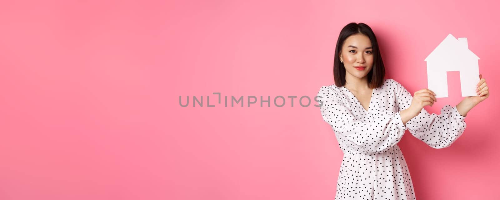 Real estate. Beautiful asian woman demonstrating paper house model, looking at camera confident, advertising home for sale, standing over pink background.