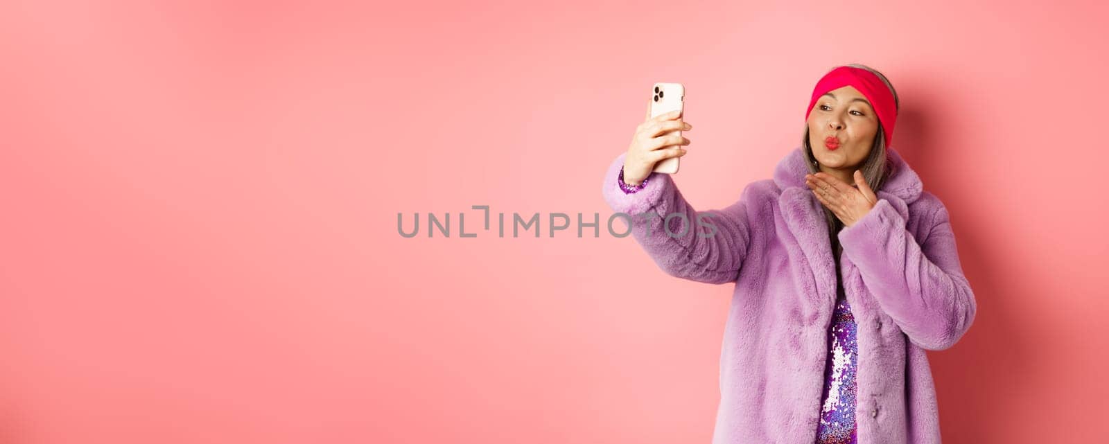 Fashion concept. Beautiful and trendy asian mature woman in faux fur coat with party dress, taking selfie, sending kiss to smartphone camera, posing against pink background by Benzoix