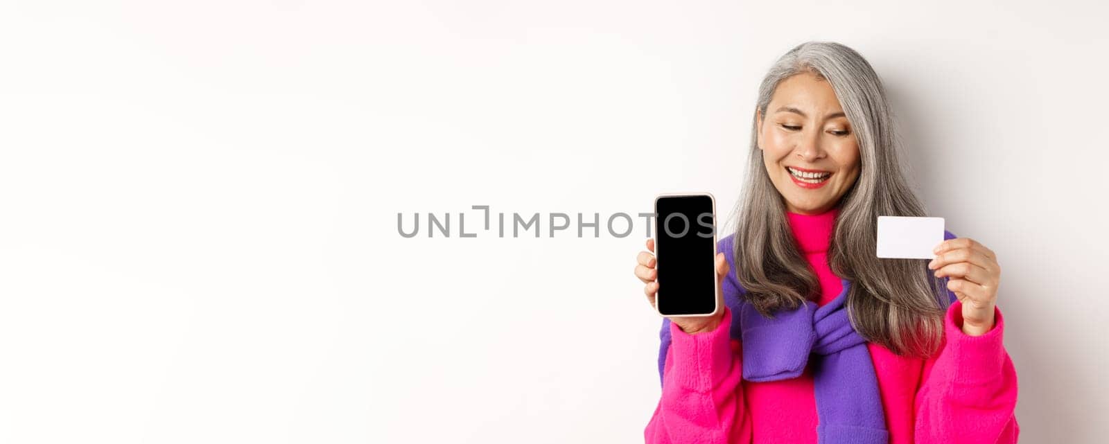 Online shopping. Closeup of fashionable old woman showing blank smartphone screen, looking pleased at plastic credit card, standing over white background.