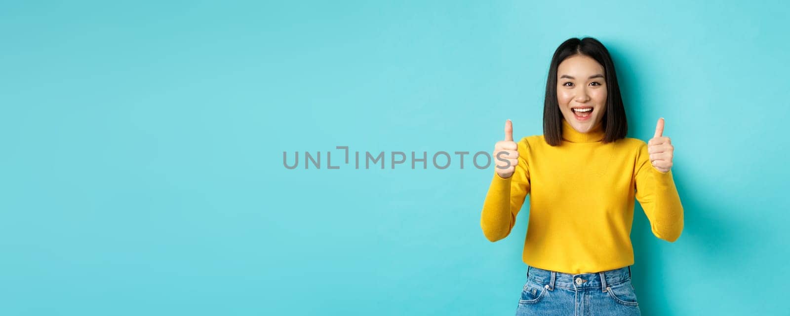 Beautiful asian woman praise good work, showing thumbs up gesture and smiling in approval, recommend product, standing satisfied over blue background.