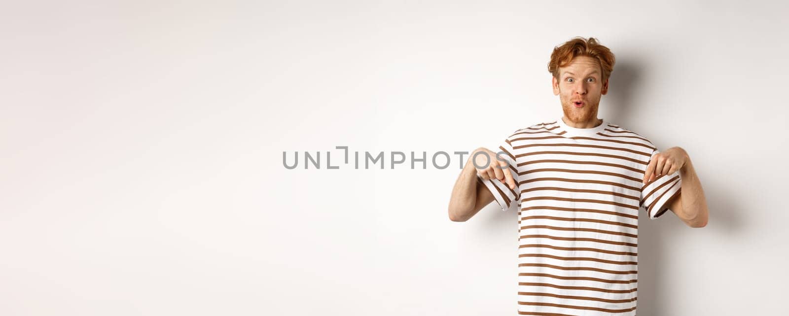 Image of funny redhead male student pointing fingers down, showing promo offer with excited smile, standing over white background.