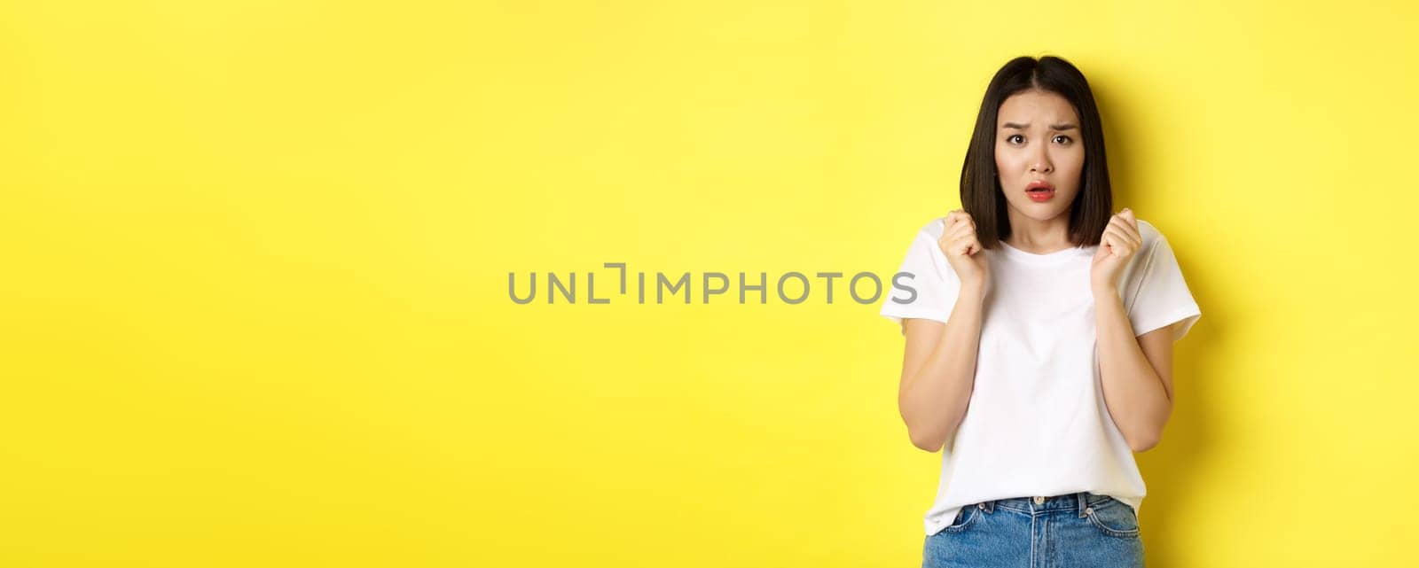 Beauty and fashion concept. Scared timid asian girl looking anxious, gasping startled and standing over yellow background.