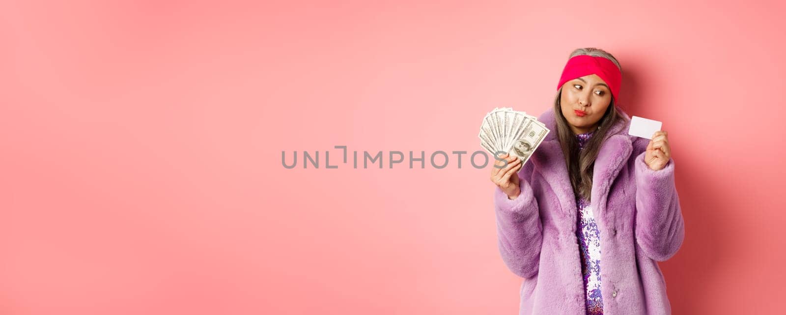 Shopping and fashion concept. Thoughtful asian female holding dollars but thinking about paying contactless with plastic credit card by Benzoix