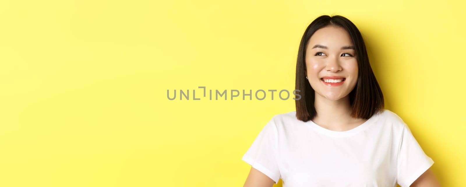 Close up of good-looking asian woman in white t-shirt, smiling and glancing left with pleased face, standing over yellow background.