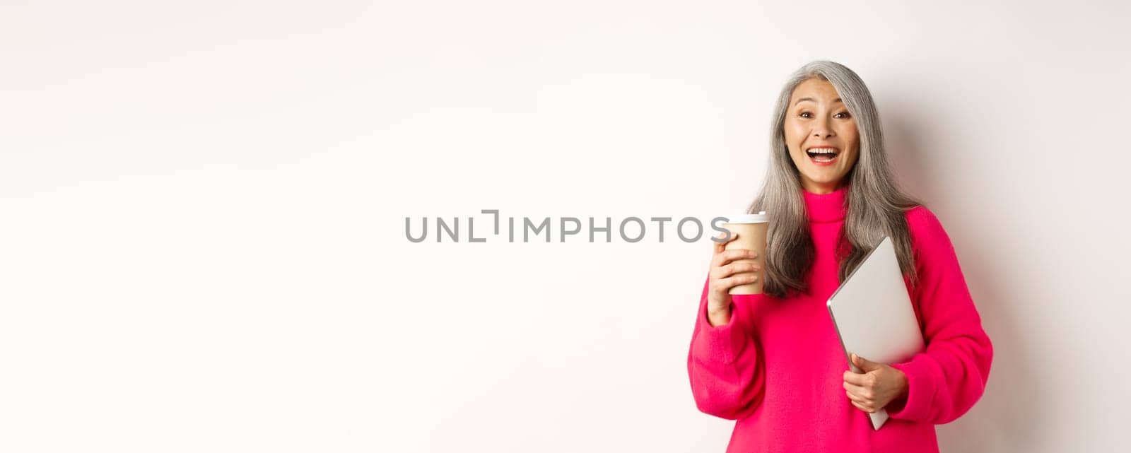 Happy asian senior female entrepreneur laughing, drinking coffee and holding laptop, standing in pink sweater over white background by Benzoix