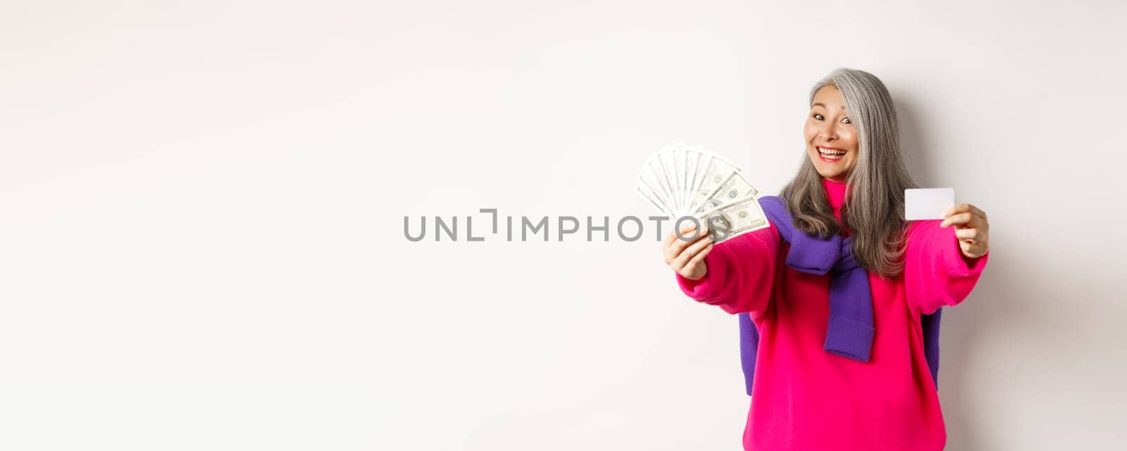 Shopping concept. Cheerful asian senior woman showing cash and plastic credit card, smiling at camera, standing over white background by Benzoix