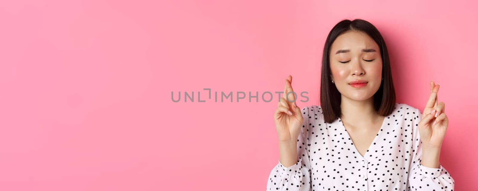 Beauty and lifestyle concept. Close-up of cute asian girl making a wish, close eyes and cross fingers hopeful, praying and standing over pink background by Benzoix