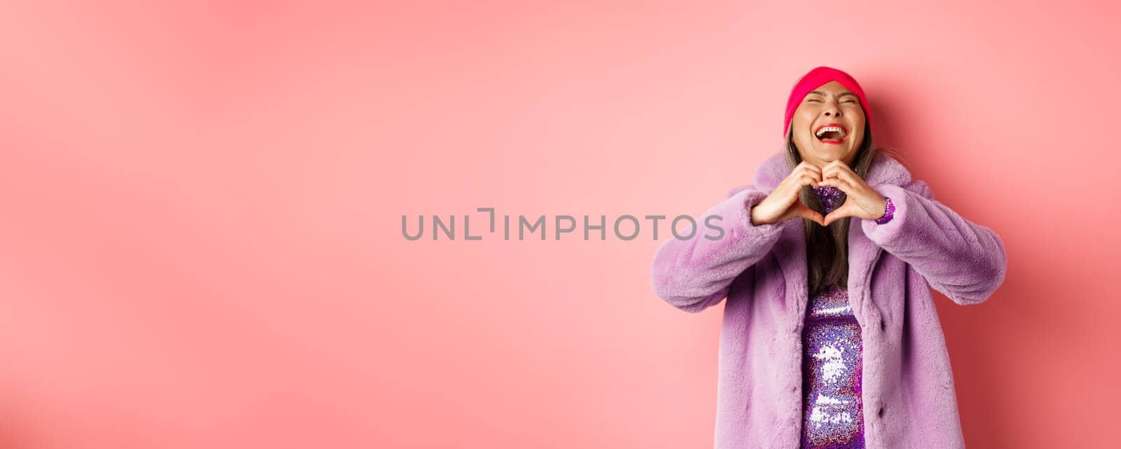 Valentines day and shopping concept. Happy asian woman in stylish outfit showing heart sign and laughing carefree, standing in faux fur against pink background by Benzoix