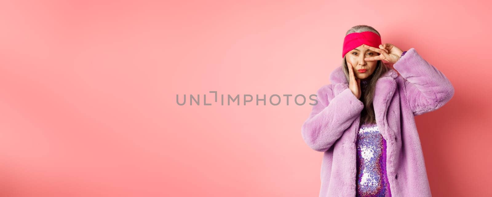 Fashion and shopping. Beautiful asian senior woman in stylish faux fur coat and headband, making peace sign on face and looking sassy and confident at camera, pink background.