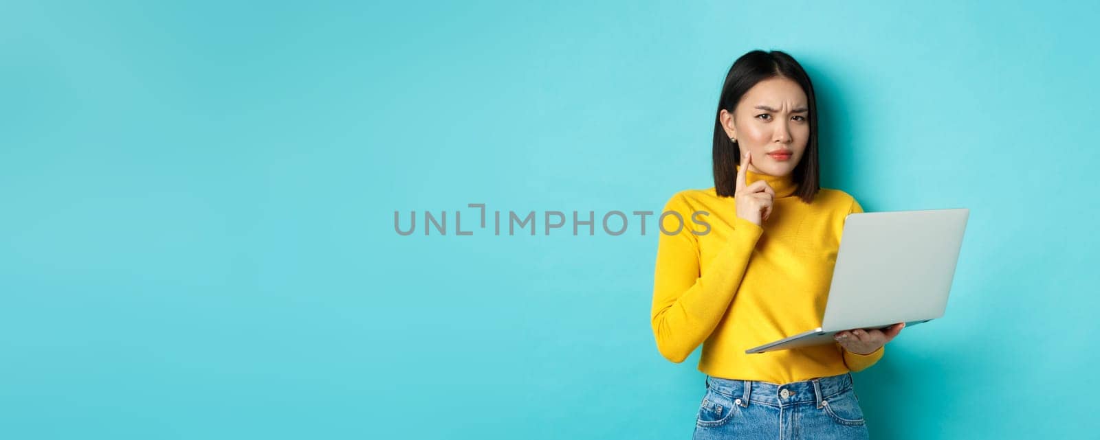 Serious looking asian woman working on laptop and thinking, frowning at camera, solving problem at work, standing over blue background.