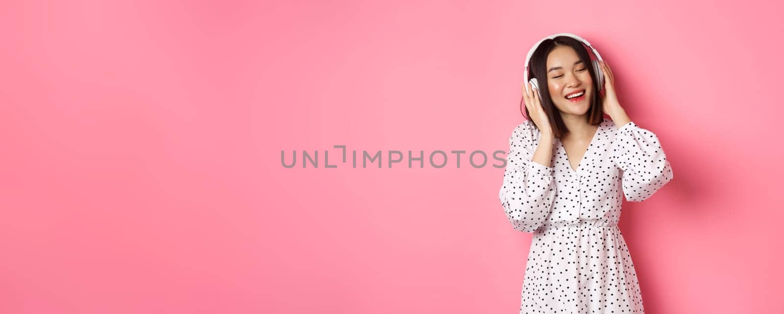 Romantic asian woman smiling happy, listening music in headphones and dancing, standing in trendy dress over pink background.